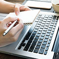 Laptop Tastatur und zwei Hände die einen kleinen Block und einen Stift halten