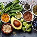 Viele kleine Schüsseln mit verschiedenen Samen nebeneinander sowie eine Avocado und Blattgemüse
