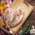 Ein Stück gewürztes helles Fleisch liegt auf einem Holzbrett umgeben von Kräutern und Öl