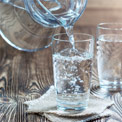 Zwei Wassergläser stehen auf einem hölzernen Untergrund und in eines wird aus einem Glaskrug Wasser eingeschenkt
