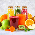 Zwei Gläser mit rotem und orangem Smoothie und dahinter drei Flaschen gefüllt mit Smoothies umgeben von Obst